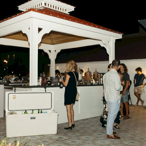 A group of people gather around an outdoor bar under a gazebo at night. The setting appears to be casual and social.