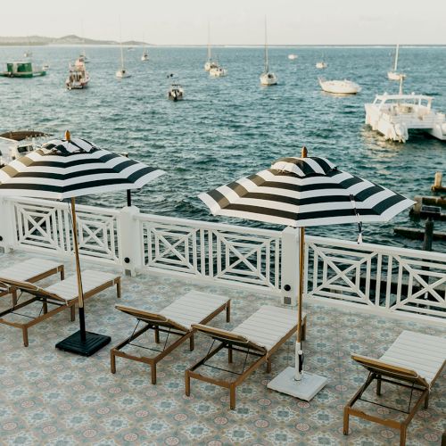 Poolside scene with striped umbrellas, lounge chairs, and boats on a calm sea under a clear sky, creating a serene coastal vibe.