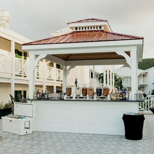 An outdoor bar setup under a gazebo with drink dispensers and coolers, set on a tiled patio near a building.