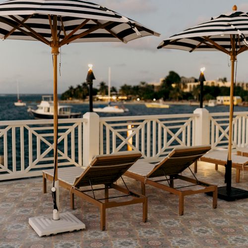 Beach loungers with striped umbrellas face the ocean. A scenic view of boats and an island is visible in the background.
