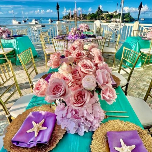 A seaside table setup features pink roses, turquoise tablecloth, purple napkins with starfish, gold chairs, and a beautiful ocean view.