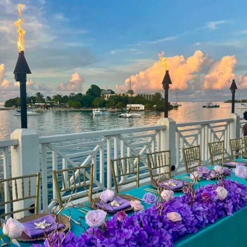A beautifully set table with purple flowers overlooks a scenic waterfront under a blue sky with torches lit along the railing.