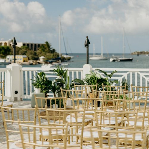 An outdoor event setup with chairs and plants overlooking a waterfront with boats and buildings in the background.