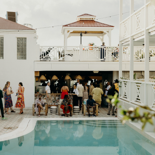 A gathering by a pool with people socializing, colorful attire, and a two-level structure with a gazebo and string lights above.