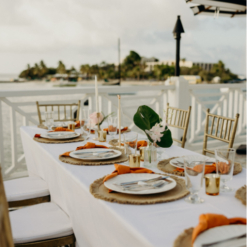 An elegant outdoor dining setup with a white tablecloth, decorated plates, orange napkins, and gold chairs, overlooking a scenic view.