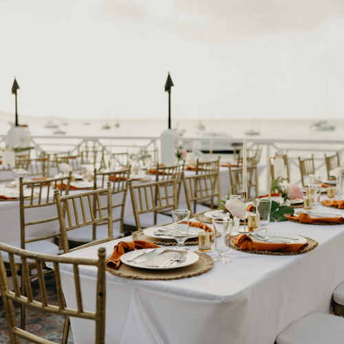 An outdoor event setup with white tablecloths, gold chairs, and nautical backdrop. Tables are decorated with plates, glasses, and flowers.