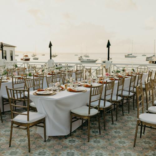 An elegant outdoor dining setup with decorated tables and chairs overlooking a serene ocean with anchored boats, under a soft sunset sky.