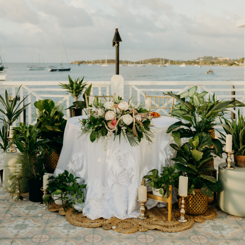 A beautifully decorated table with a white cloth, flowers, and surrounded by greenery, set against a stunning waterfront view.