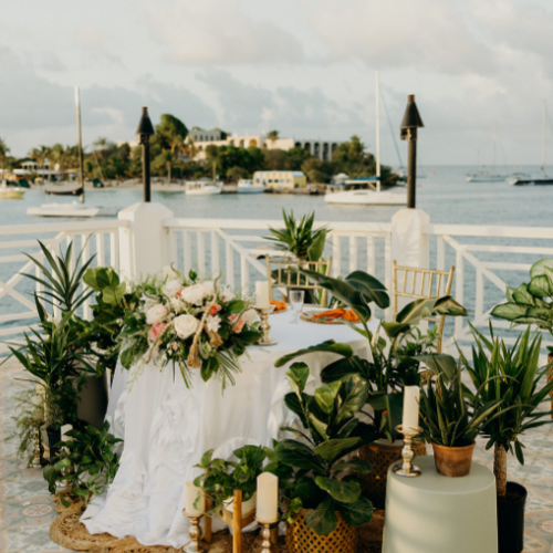 A beautifully decorated outdoor setup with a floral arrangement, green plants, candles, and a view of the sea with boats in the background.