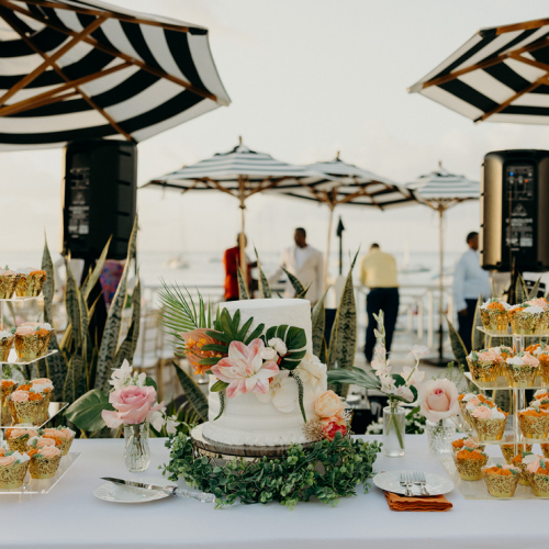 A decorated cake with floral elements is on a table surrounded by cupcakes under striped umbrellas, set for an outdoor event.