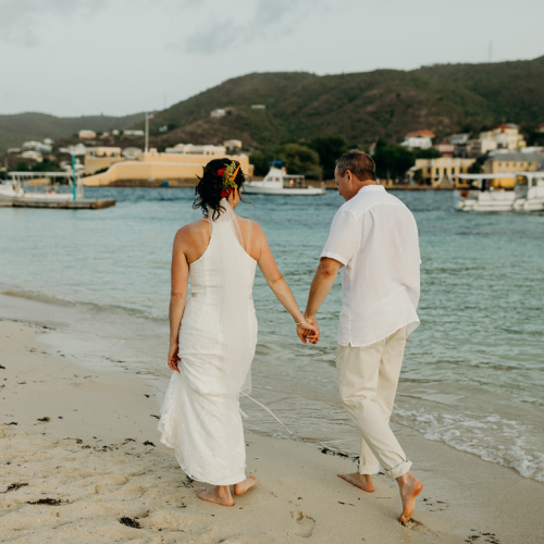 A couple in white attire holds hands while walking on a beach beside calm waters and distant hills.
