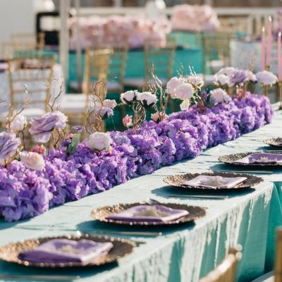 A table is elegantly set with turquoise cloths, lilac napkins, gold-rimmed plates, and lavish purple floral arrangements.