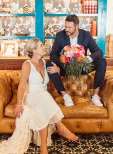 A bride in a white dress sits on a tan leather couch while a groom in a navy suit hands her a bouquet of pink, orange, and peach flowers.