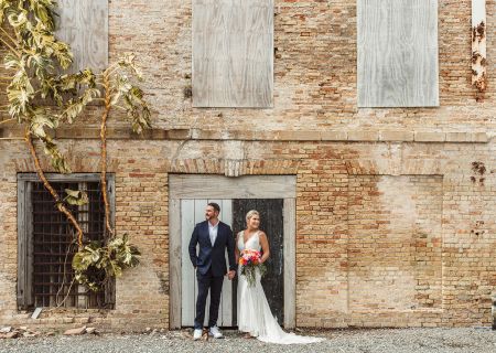 A wedding party stands before a rustic stone building, the couple in the doorway with guests gathered around, outdoors by warm sunlight.
