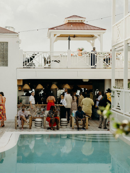 A lively outdoor gathering by a pool, with people socializing near a two-story building under a gazebo.