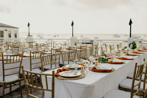 Outdoor wedding reception setup with white tablecloths, gold chairs, floral arrangements, and ocean view in background.