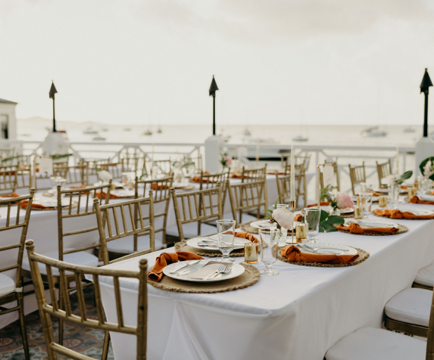 A long outdoor dining setup with many wooden tables and chairs, white plates, and warm string lights under a light-filled sky, styled for a wedding or event.