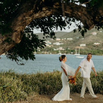 A couple is walking hand in hand near a scenic waterfront, surrounded by lush greenery and distant hills.