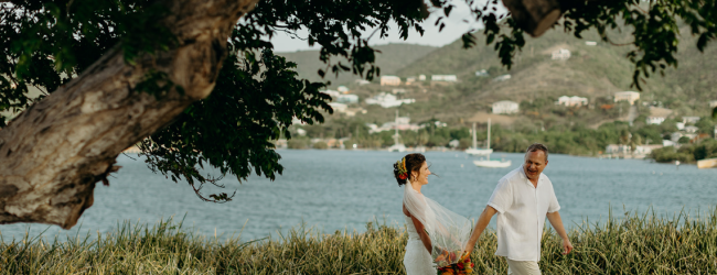 A couple holding hands and dancing in a rustic outdoor setting by a lake, with trees and a wooden arch, celebrating joy.