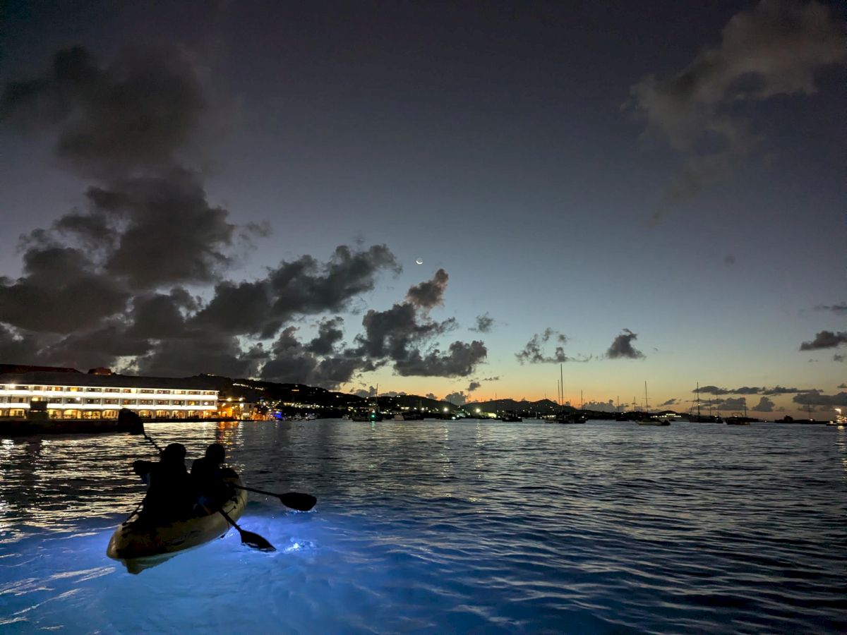 Kayak on a calm harbor at dusk, boats and lights along the shore as stormy clouds frame a pale moon.