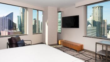A modern hotel room with city views from large windows, featuring a bed, TV, armchair, and table.