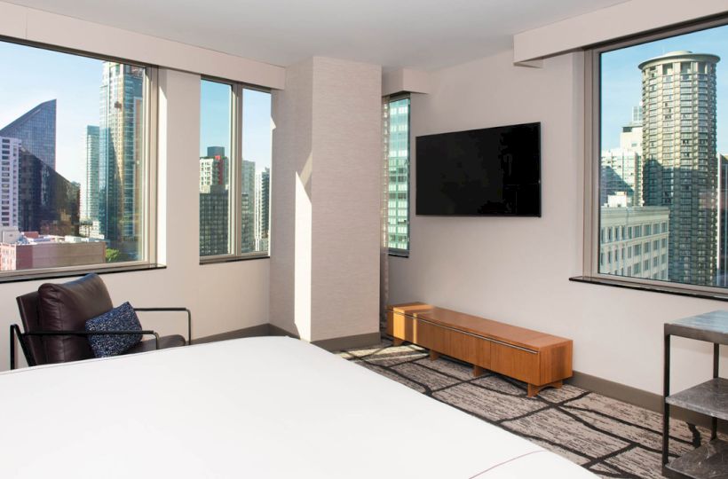A modern hotel room with city views from large windows, featuring a bed, TV, armchair, and table.