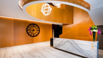 A modern interior lobby with wooden accents, a large clock, stylish lighting, and a marble reception desk with flowers on top.
