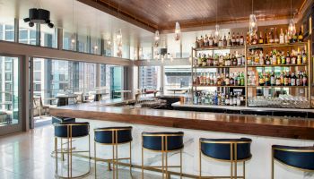A modern bar with a stocked shelf, five bar stools, and large windows overlooking a cityscape. Hanging lights add a warm ambiance.