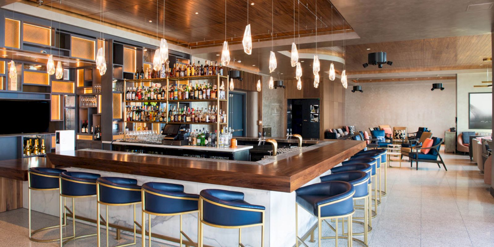 A modern bar with a stocked shelf, stylish lighting, and blue cushioned stools around a wooden counter in a spacious lounge area.