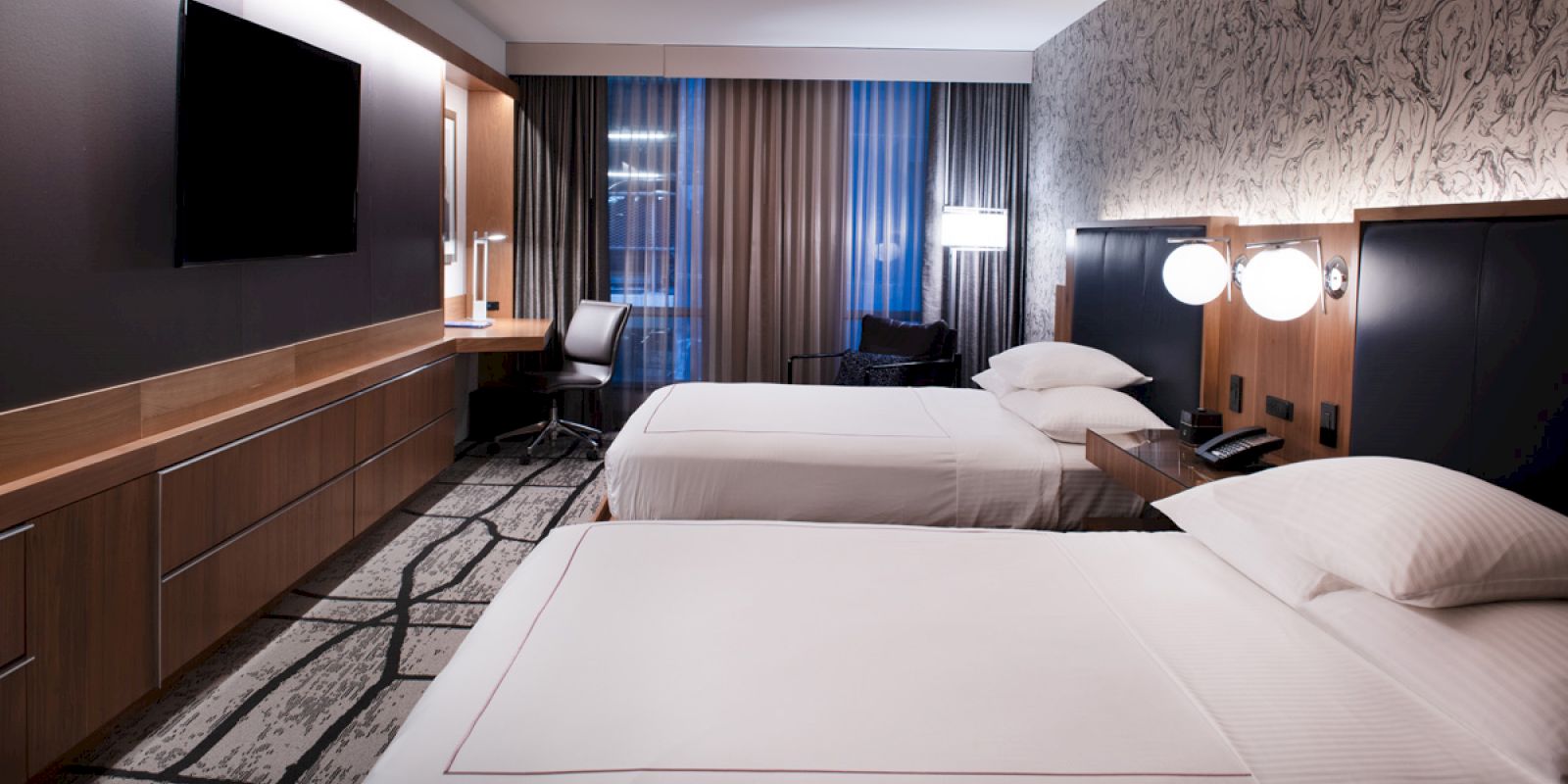 A modern hotel room with two beds, a wall-mounted TV, desk area, and trendy lighting. Neutral tones and a patterned carpet complete the look.