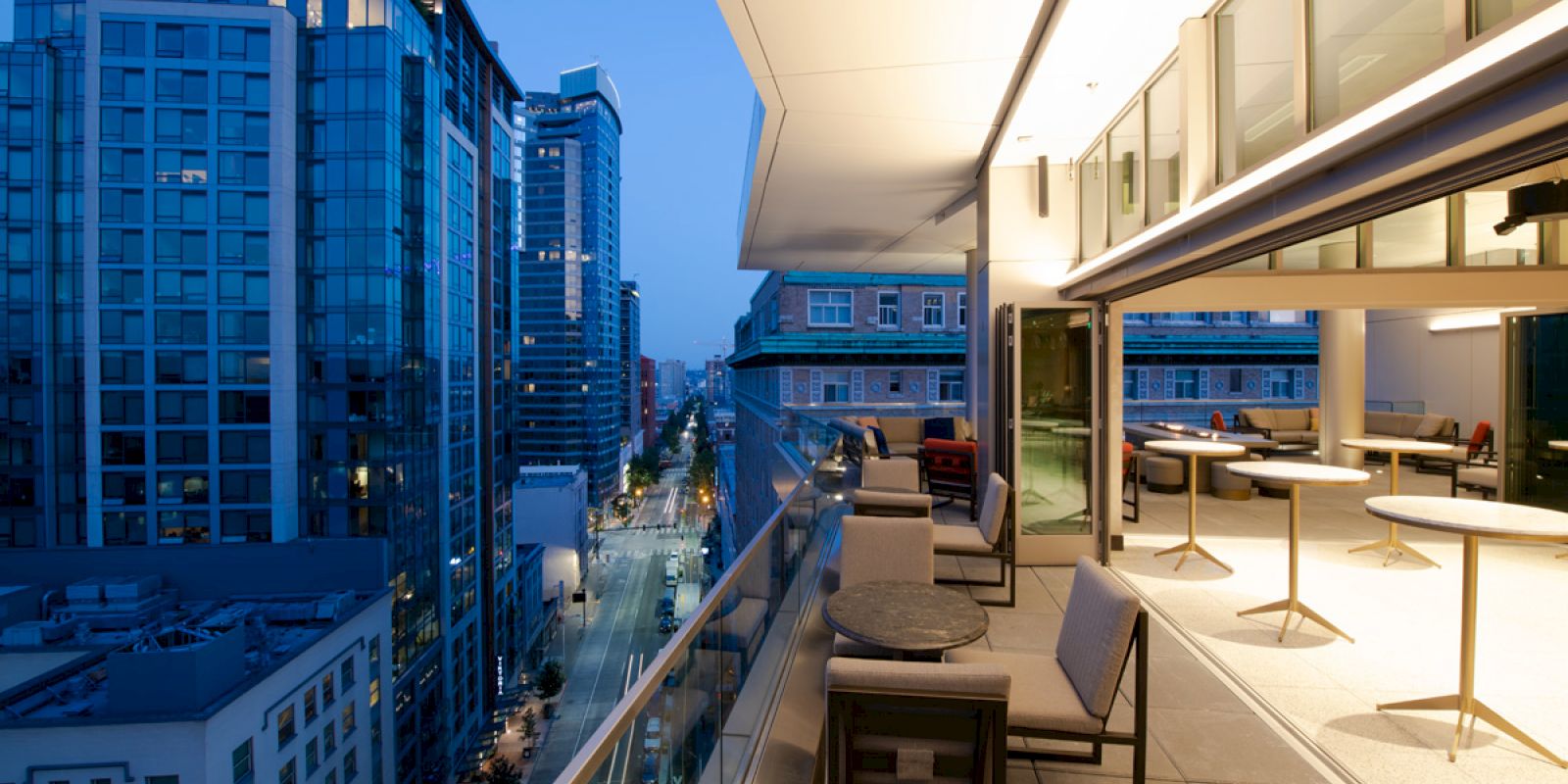 A modern cityscape with high-rise buildings, a street view, and a chic balcony featuring seating and tables, captured during twilight.