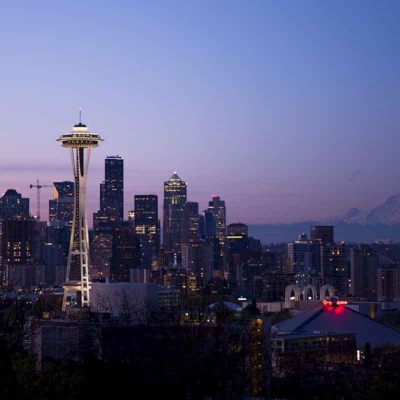 The image shows a city skyline at dusk featuring the Space Needle, with Mount Rainier in the background and city lights beginning to illuminate.