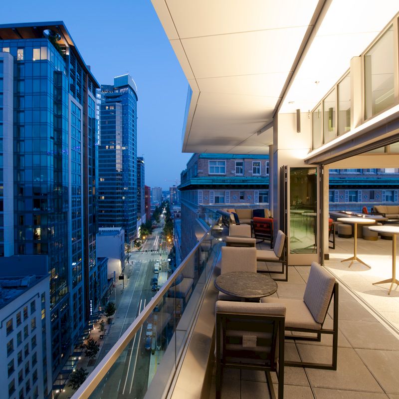 A modern balcony overlooking a city street, featuring comfortable seating and tables, with buildings and lights in the evening sky.