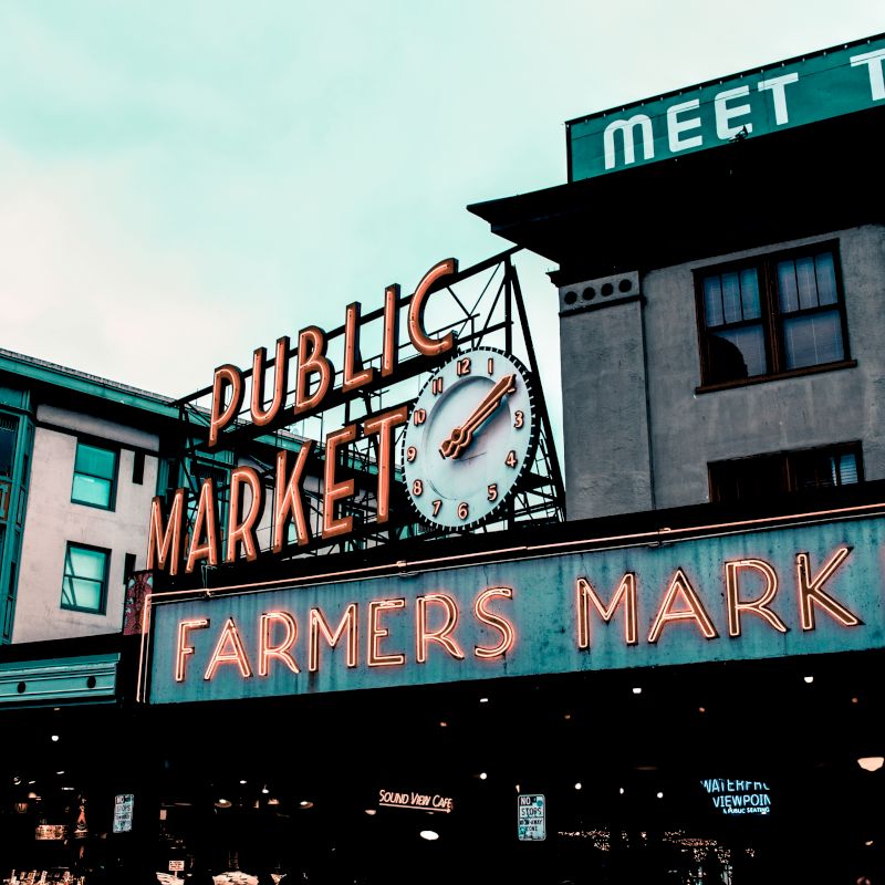 A public market sign with a clock and a neon "Farmers Market" sign, set against old-style buildings under a cloudy sky.