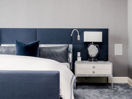 A modern bedroom features a navy-blue headboard, white lamp on a nightstand, and a neatly made bed with gray and blue accents.