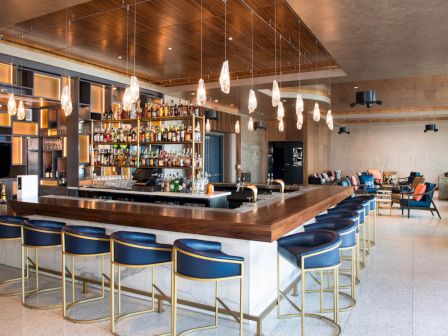 A modern bar with blue stools, hanging lights, and a stocked liquor shelf; cozy seating area in the corner of a spacious, stylish room.
