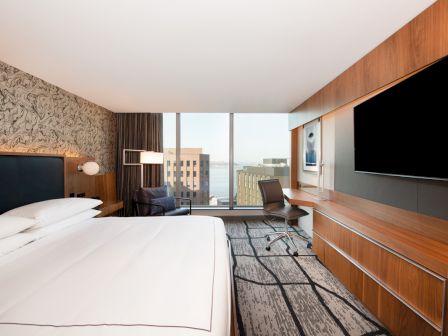 A modern hotel room with a large bed, wall TV, desk, and floor-to-ceiling windows overlooking a city view.