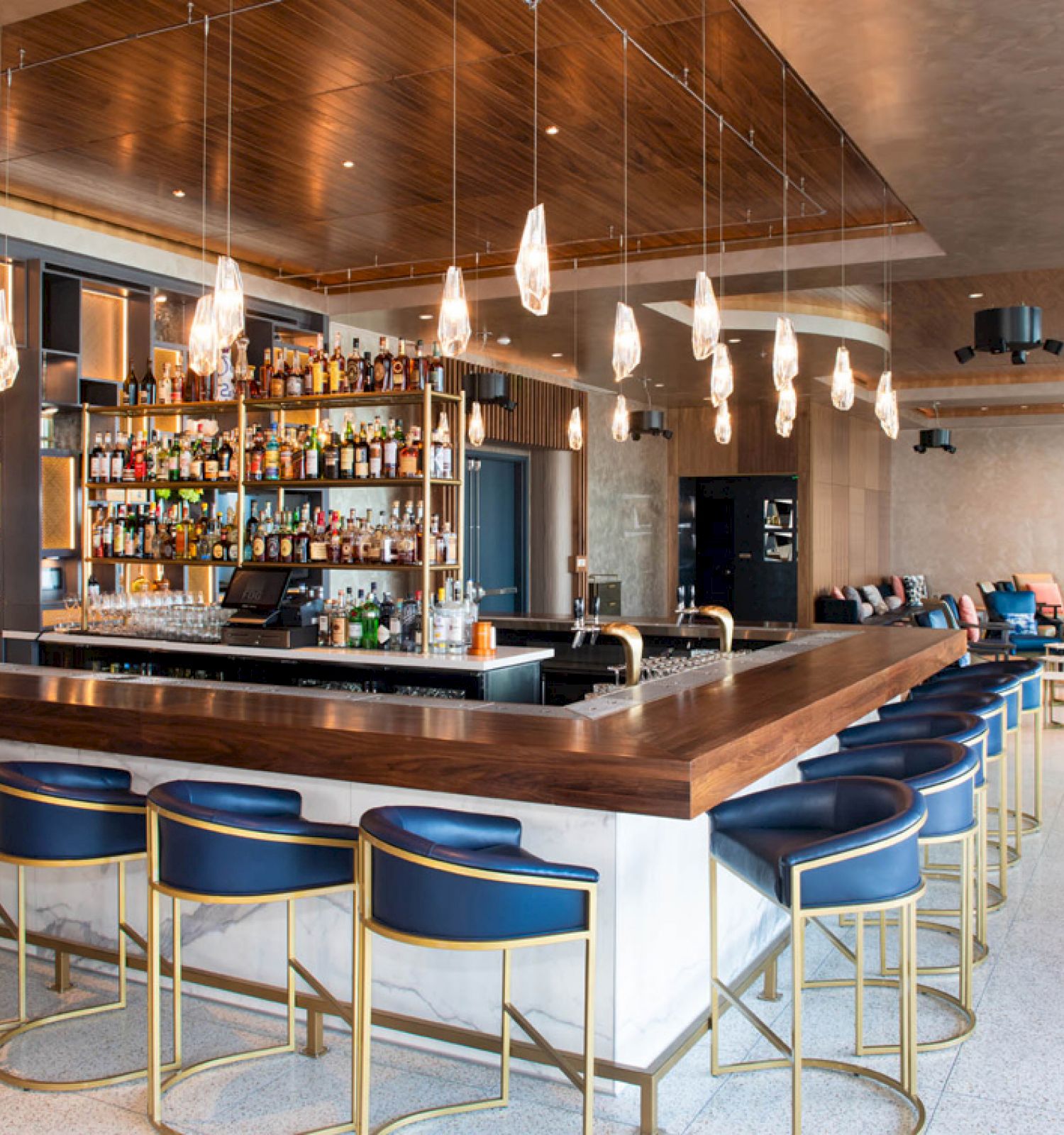 The image shows a stylish, modern bar with blue stools, a central bar counter, shelves of liquor, and pendant lights.