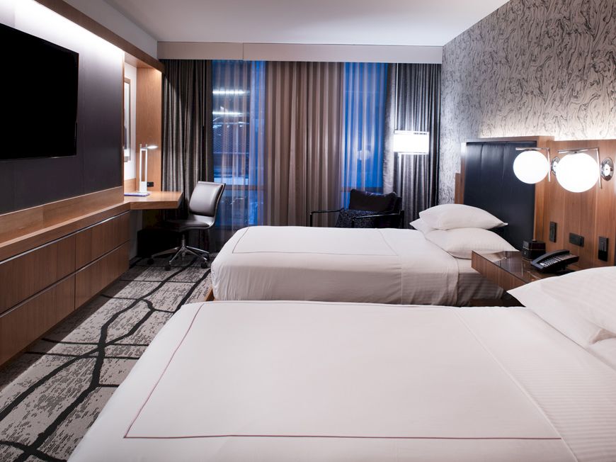 The image shows a modern hotel room with two beds, a desk, large TV, and stylish decor with dark curtains and patterned carpet.