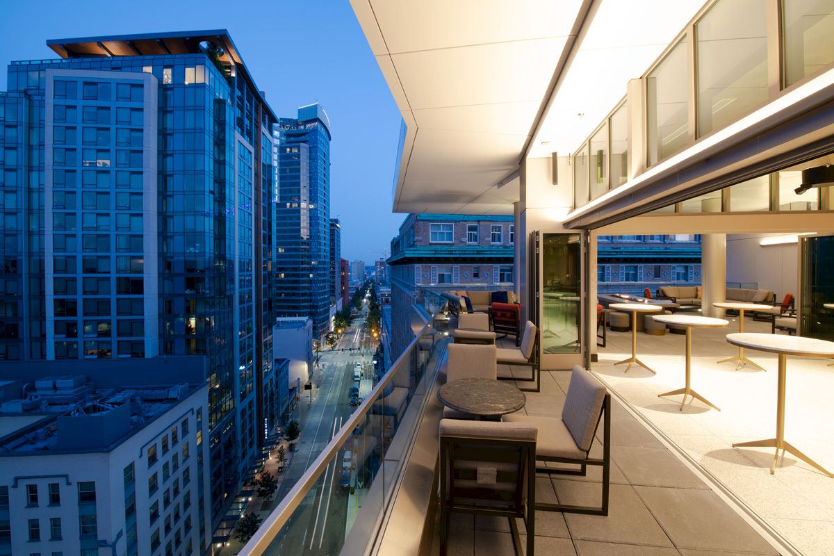 A cityscape viewed from a modern balcony with tables and chairs, showcasing tall buildings and a street below in the early evening.