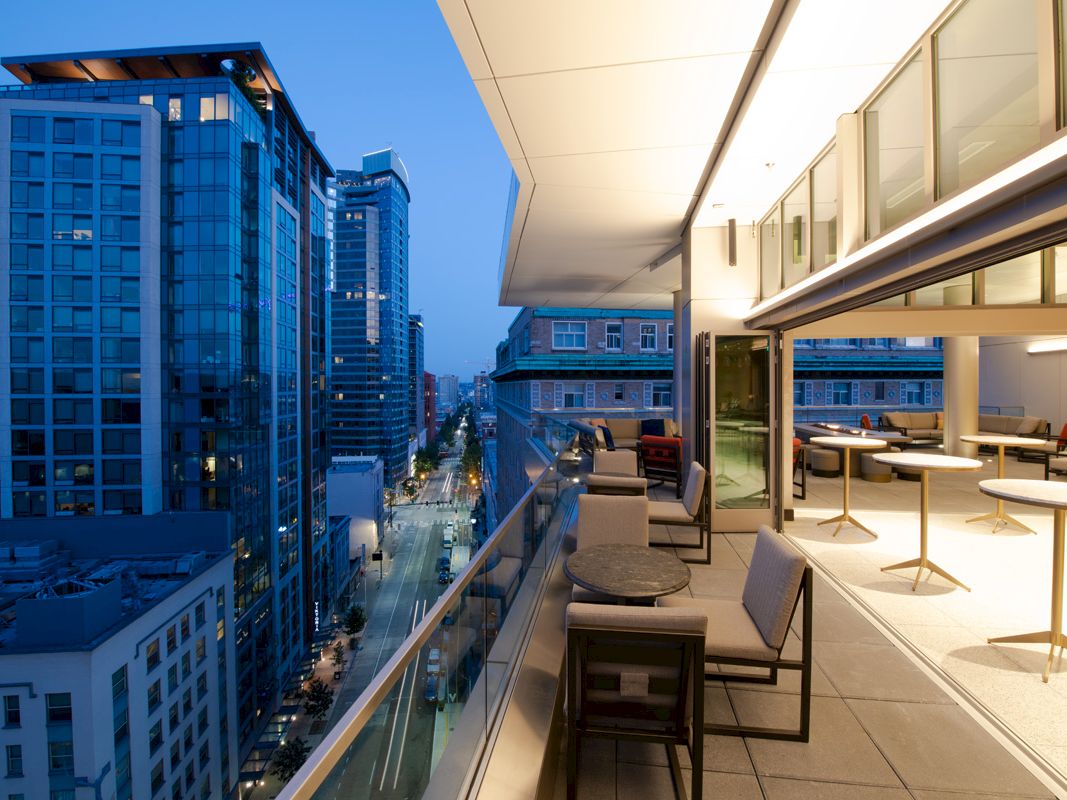 A cityscape viewed from a modern balcony with tables and chairs, showcasing tall buildings and a street below in the early evening.