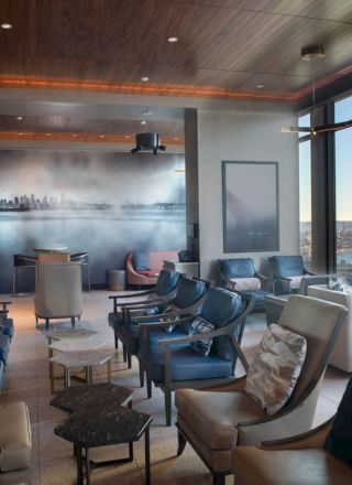 A modern, stylish lounge with leather chairs, large windows overlooking water, and skyline wall art. Natural light fills the spacious room.