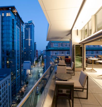 A modern rooftop lounge overlooks a city street with tall buildings, featuring seating areas and large windows at dusk.