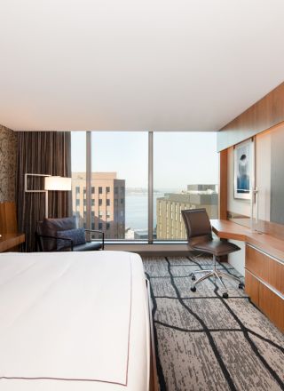 A modern hotel room with a large bed, flat-screen TV, desk, and window view of buildings and water.
