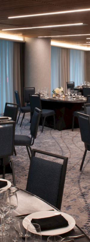 The image shows an elegantly set dining room with round tables, black chairs, and floral centerpieces in a modern, dimly lit space.