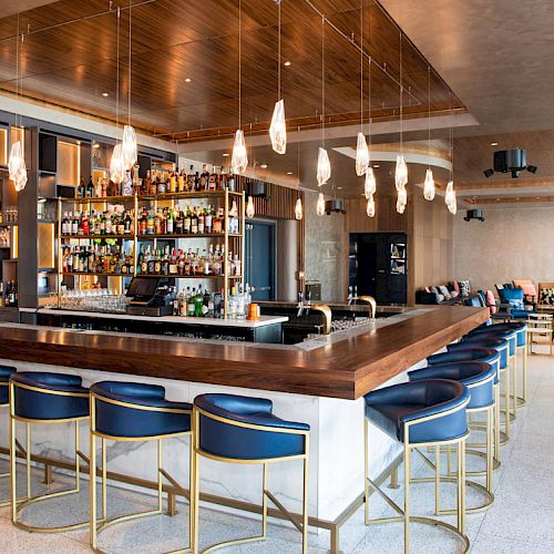 A modern bar with blue chairs, pendant lights, and a variety of spirits on shelves. There's cozy seating in the background.