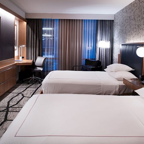 The image shows a hotel room with two beds, a TV on the wall, a desk with a chair, and modern lighting, featuring a patterned carpet and wallpaper.