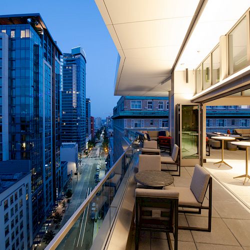 A modern cityscape at dusk, featuring a stylish rooftop terrace with seating, overlooking tall buildings and a lit-up street below.