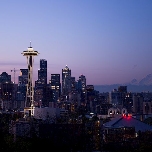 The image shows a view of Seattle's skyline at dusk, featuring the Space Needle, skyscrapers, and Mount Rainier in the background.