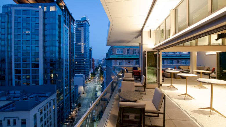 A modern rooftop patio with tables and chairs overlooks a cityscape at dusk, featuring tall buildings with illuminated windows.